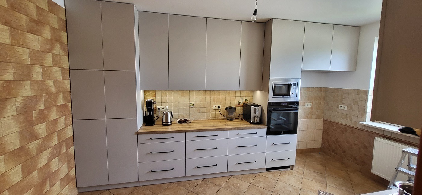 Jasnoszara zabudowa kuchenna z drewnianym blatem, oświetleniem podszafkowym i czarnymi uchwytami szuflad, widok na ścianę z cegiełkową tapetą