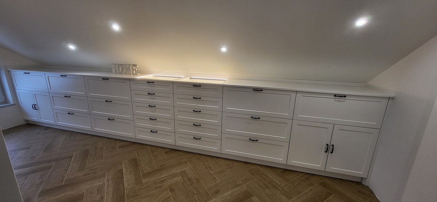 Białe szafki i szuflady z czarnymi uchwytami, zabudowane pod skosem na poddaszu, z drewnianą podłogą w jodełkę.