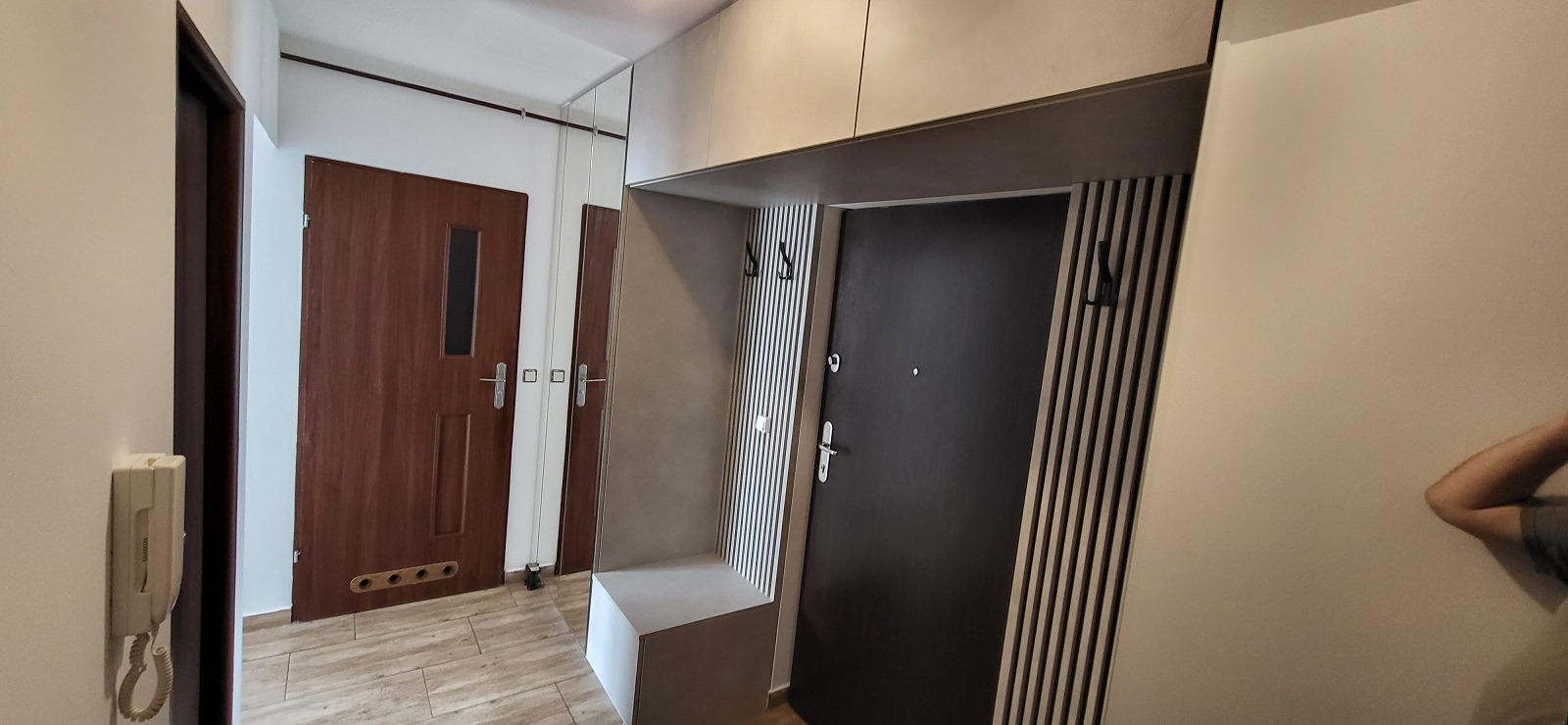 Wnętrze przedpokoju z zabudową meblową obejmującą szafę z wieszakami, siedzisko oraz szafki górne, z drzwiami wejściowymi obok i drzwiami wewnętrznymi po lewej stronie.