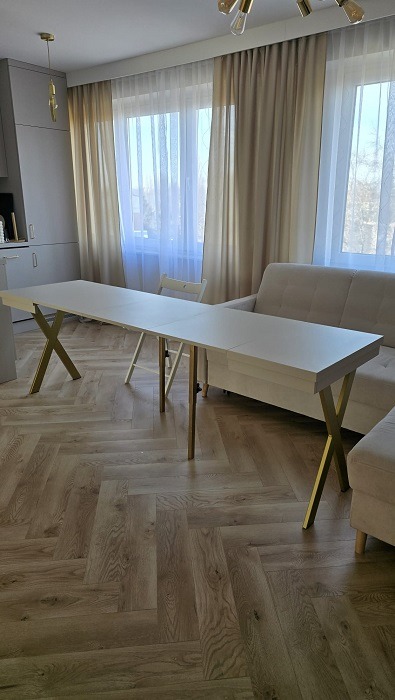 Rozkładany stół z białym blatem i złotymi nogami w kształcie litery X, ustawiony w salonie z parkietem w jodełkę i beżową sofą.