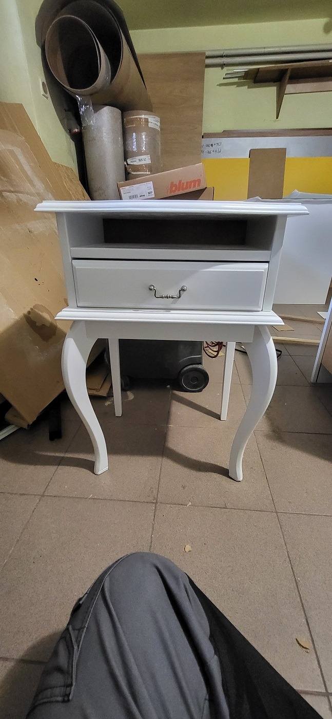 Biała szafka nocna z szufladą i giętymi nogami, sfotografowana w warsztacie w Lublinie, z widocznymi materiałami stolarskimi w tle.