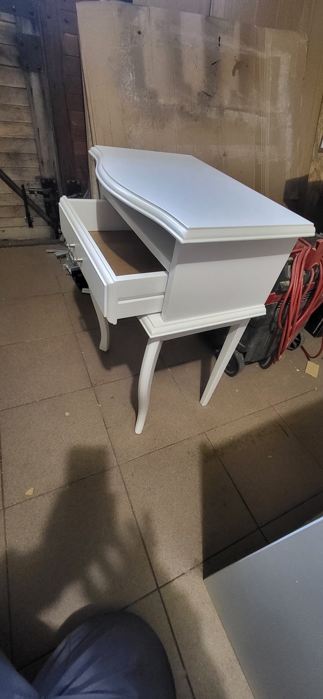 Biała toaletka w stylu Ludwika z wysuniętą szufladą, widoczne gięte nogi i frezowane krawędzie, tło warsztatowe.