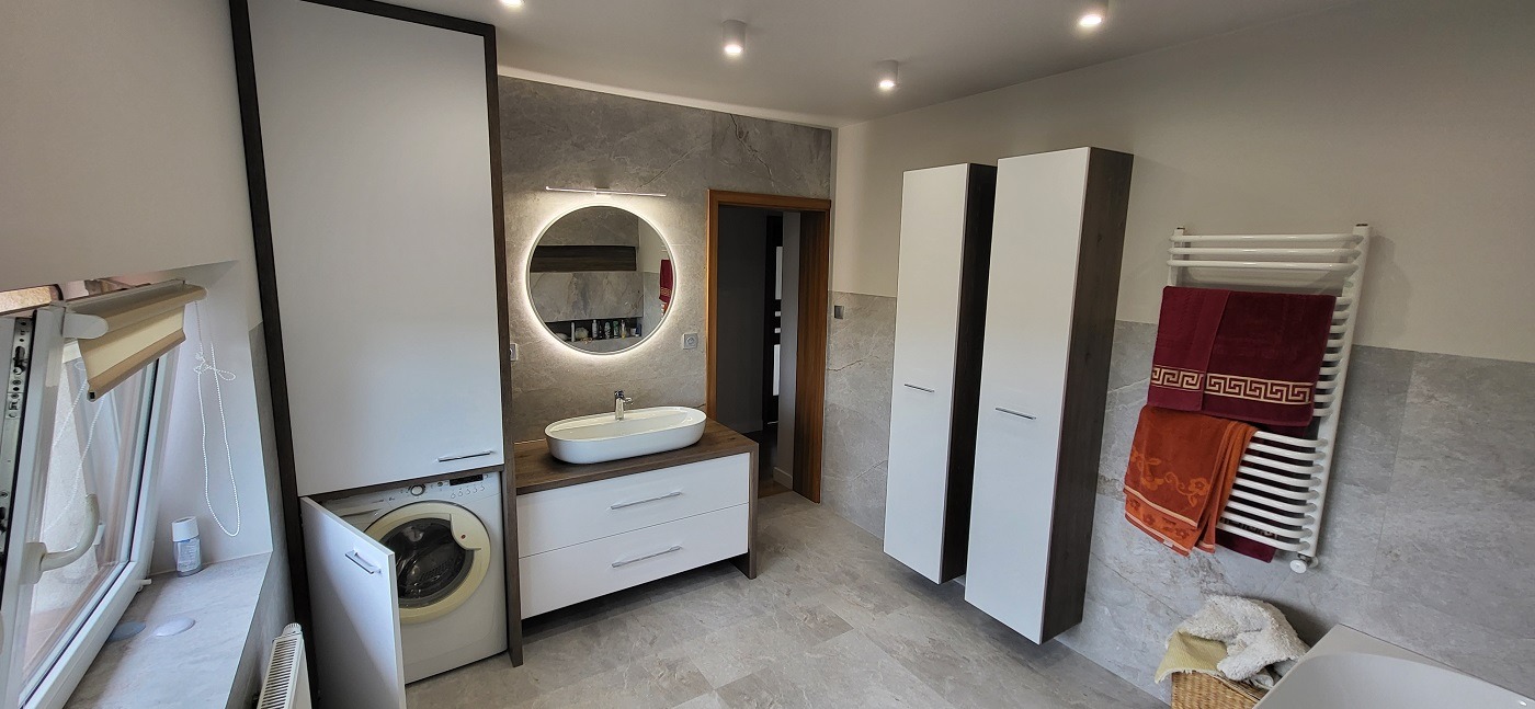 Nowoczesna łazienka z pralką zabudowaną w szafce, okrągłym lustrem z podświetleniem LED, umywalką nablatową, grzejnikiem drabinkowym z ręcznikami i wysokimi szafkami.