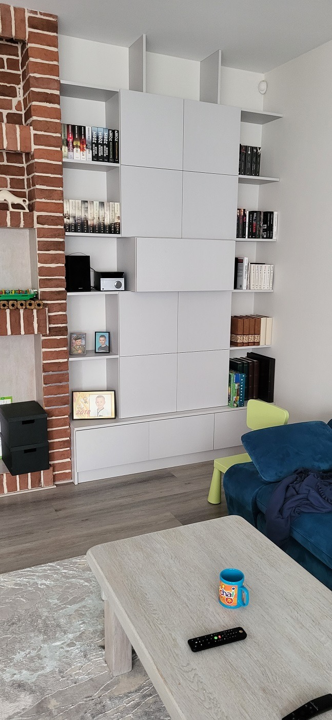 Biała zabudowa meblowa na ścianie z półkami na książki, elementem dekoracyjnym z cegły i widocznym fragmentem salonu z niebieską kanapą oraz stolikiem kawowym z pilotem i kubkiem.