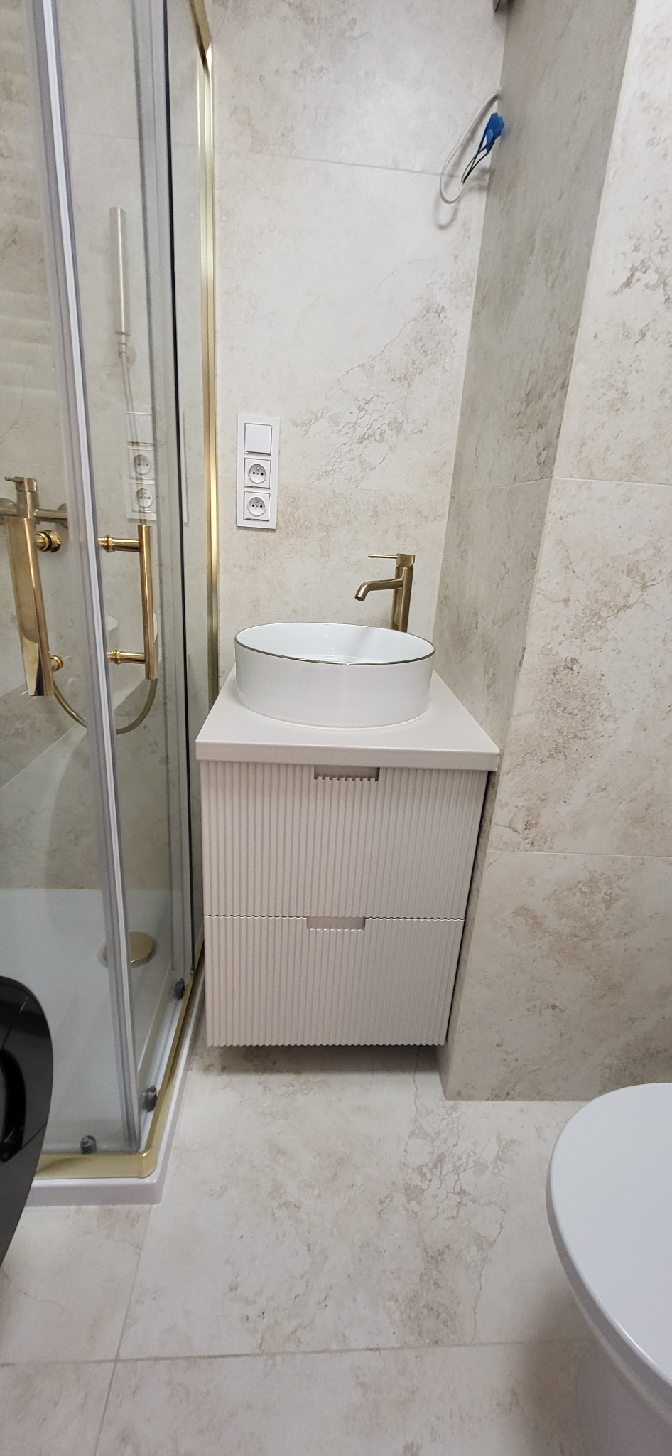 Nowoczesna łazienka z kabiną prysznicową ze złotymi detalami, umywalką nablatową z szafką w kolorze złamanej bieli i armaturą w odcieniu szczotkowanego złota.