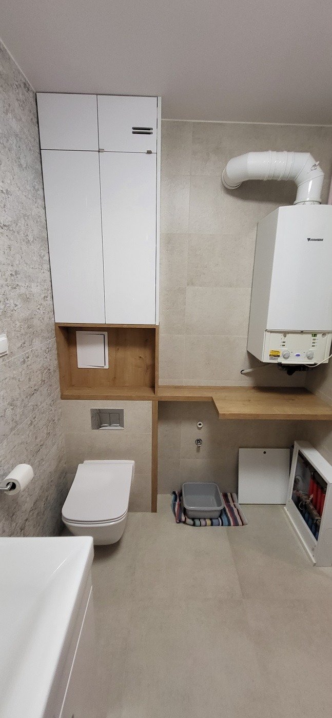 Nowoczesna łazienka z zabudową meblową z białymi frontami i drewnianymi elementami, podwieszana toaleta, piec gazowy z widoczną rurą odprowadzającą spaliny, szara kuweta na podłodze.