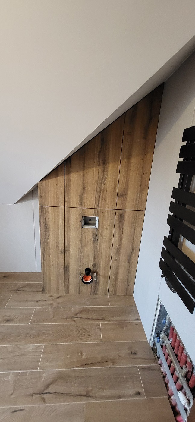 Ukryta instalacja wodno-kanalizacyjna za drewnopodobną zabudową w skośnej łazience na poddaszu, widoczne rury grzewcze i otwór rewizyjny.