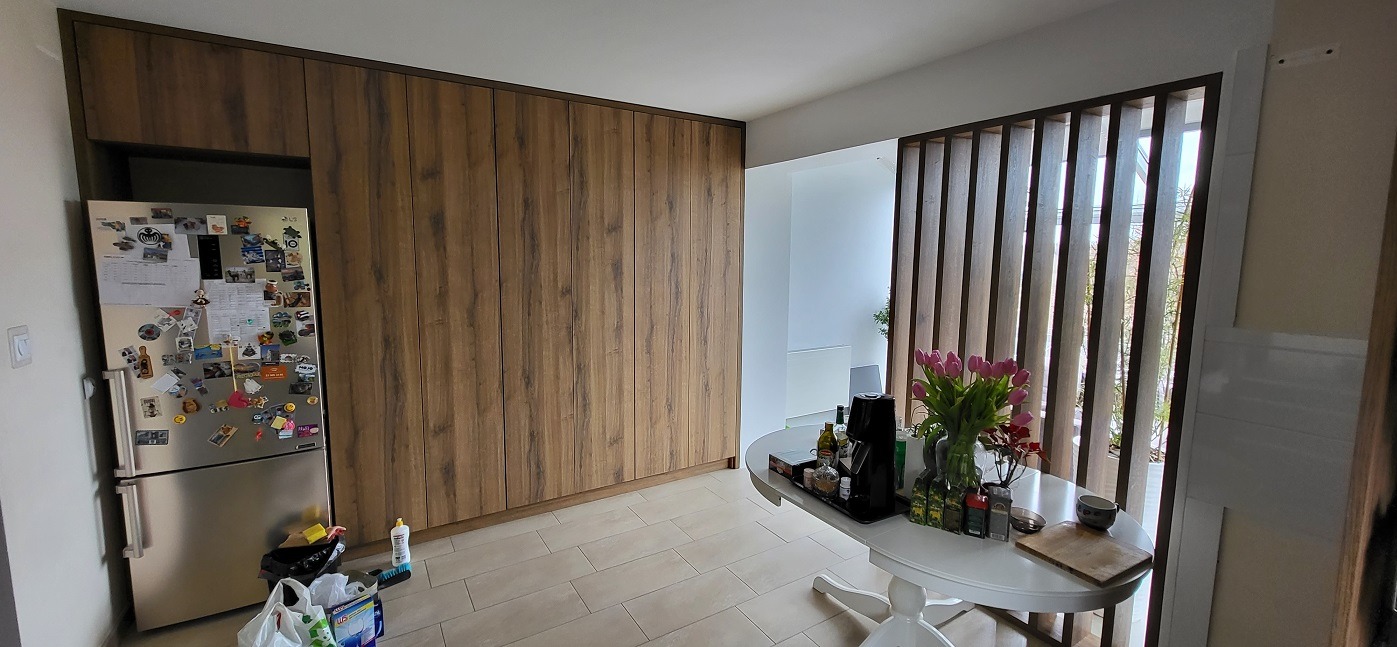 Drewniana szafa wnękowa z ukrytymi drzwiami, dopasowana do kolorystyki kuchni, z widoczną lodówką po lewej stronie i ażurową ścianką działową po prawej.