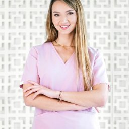 Powiększanie ust (botoks), oczyszczanie wodorowe, mezoterapia - Kosmetolog Kraków - Uśmiechnięta kobieta w różowym uniformie medycznym, ze skrzyżowanymi rękami, na tle dekoracyjnej białej ściany z geometrycznym wzorem.