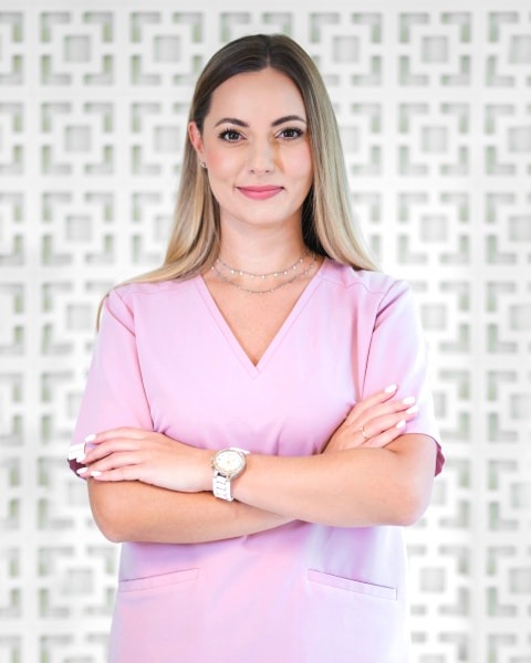 Portret kobiety w różowym uniformie medycznym, z zegarkiem na nadgarstku i złożonymi rękami na tle dekoracyjnej ściany.