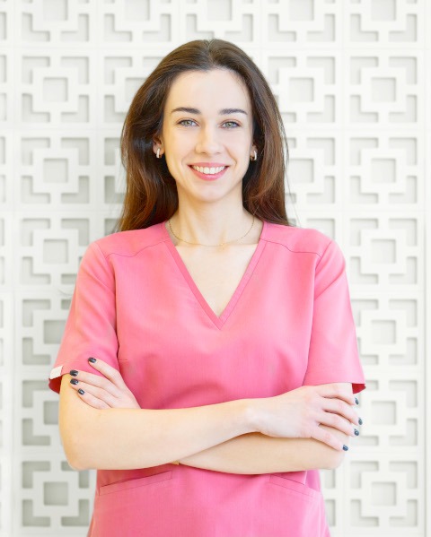 Uśmiechnięta kobieta w różowym uniformie medycznym, z rękami skrzyżowanymi na piersiach, na tle dekoracyjnej białej ściany.