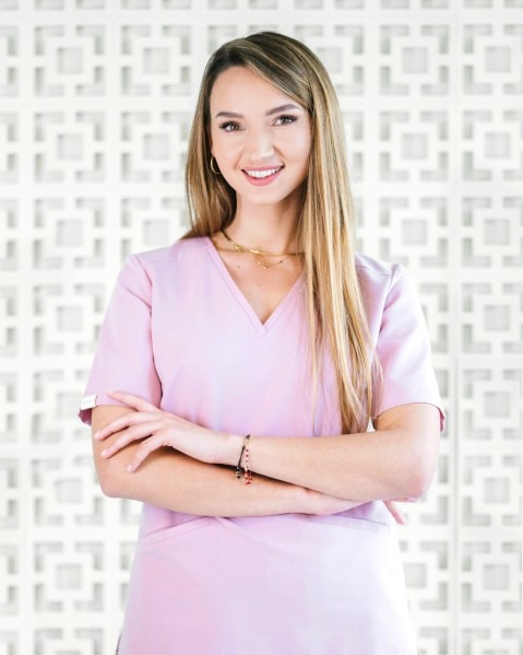 Uśmiechnięta kobieta w różowym uniformie medycznym, ze skrzyżowanymi rękami, na tle dekoracyjnej białej ściany z geometrycznym wzorem.