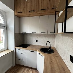 SKOTA SPÓŁKA Z OGRANICZONĄ ODPOWIEDZIALNOŚCIĄ - Nowoczesna, kompaktowa kuchnia z białymi szafkami dolnymi i drewnopodobnymi górnymi, czarnym zlewem i płytą grzewczą, minimalistyczne uchwyty, drewniana podłoga i okno z widokiem na budynek...