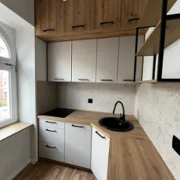 Nowoczesna, kompaktowa kuchnia z białymi szafkami dolnymi i drewnopodobnymi górnymi, czarnym zlewem i płytą grzewczą, minimalistyczne uchwyty, drewniana podłoga i okno z widokiem na budynek...