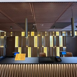 SKOTA SPÓŁKA Z OGRANICZONĄ ODPOWIEDZIALNOŚCIĄ - Recepcja hotelowa z drewnianą okładziną ścienną z podświetleniem, ciemnym blatem, kasami fiskalnymi i metalową kratką na suficie, oświetlona cylindrycznymi lampami wiszącymi.