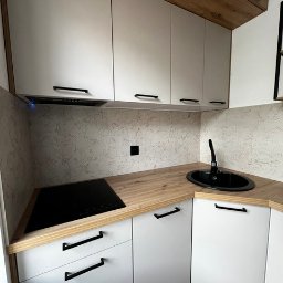 SKOTA SPÓŁKA Z OGRANICZONĄ ODPOWIEDZIALNOŚCIĄ - Nowoczesna kuchnia z białymi szafkami z czarnymi uchwytami, drewnianym blatem i czarnym zlewem owalnym, płyta indukcyjna w rogu blatu, widoczne szafki górne z drewnianym wykończeniem na górze.