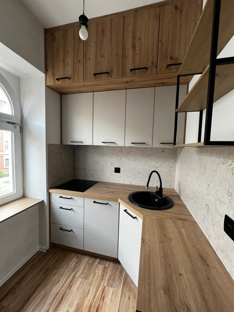 Nowoczesna, kompaktowa kuchnia z białymi szafkami dolnymi i drewnopodobnymi górnymi, czarnym zlewem i płytą grzewczą, minimalistyczne uchwyty, drewniana podłoga i okno z widokiem na budynek...
