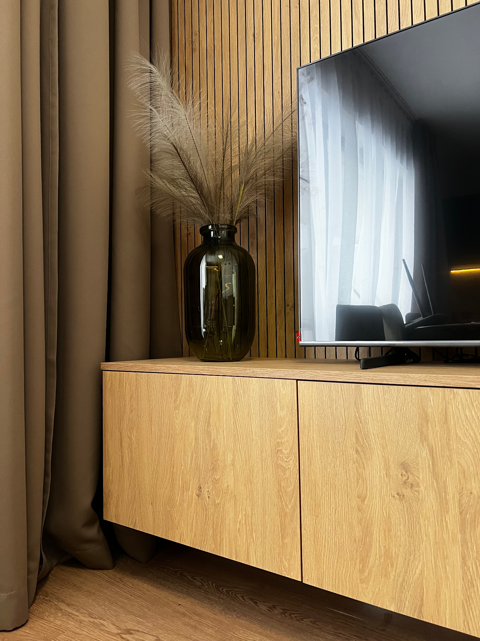 Telewizor na szafce RTV z drewnianymi frontami w salonie w Jeleniej Górze, obok zielony wazon z suszonymi trawami, tło stanowi ściana z drewnianych paneli.