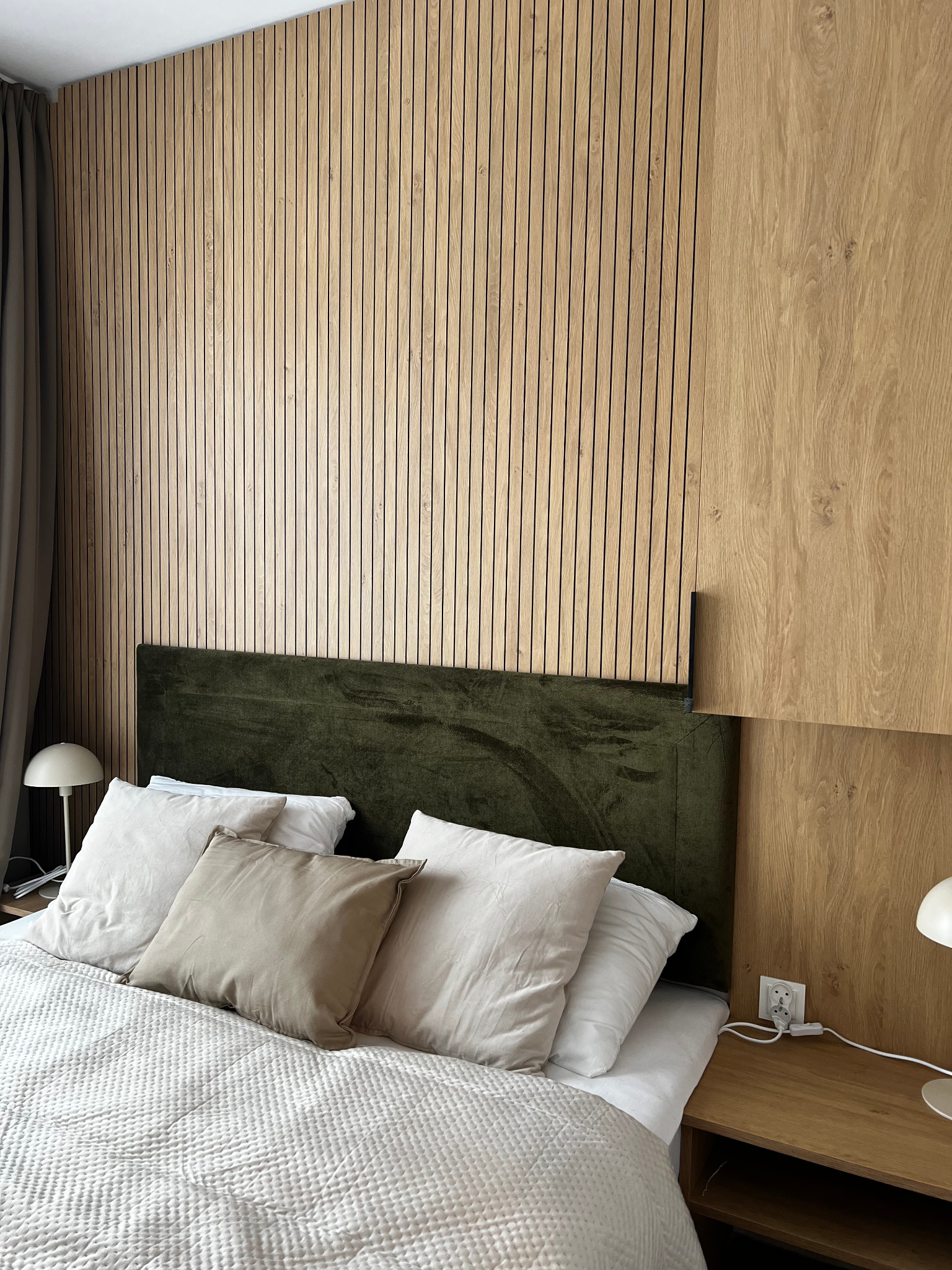 Nowoczesna sypialnia z drewnianą ścianą z paneli, zielonym zagłówkiem i beżową pościelą, oświetlona lampkami nocnymi na szafkach.