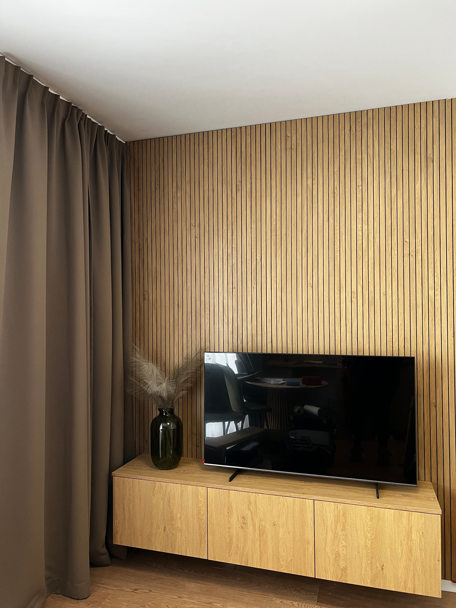 Nowoczesny salon z drewnianą ścianą z paneli, niską szafką RTV z jasnego drewna, na której stoi ciemny wazon z suchymi trawami i telewizor, zasłonięte okno brązowymi zasłonami.