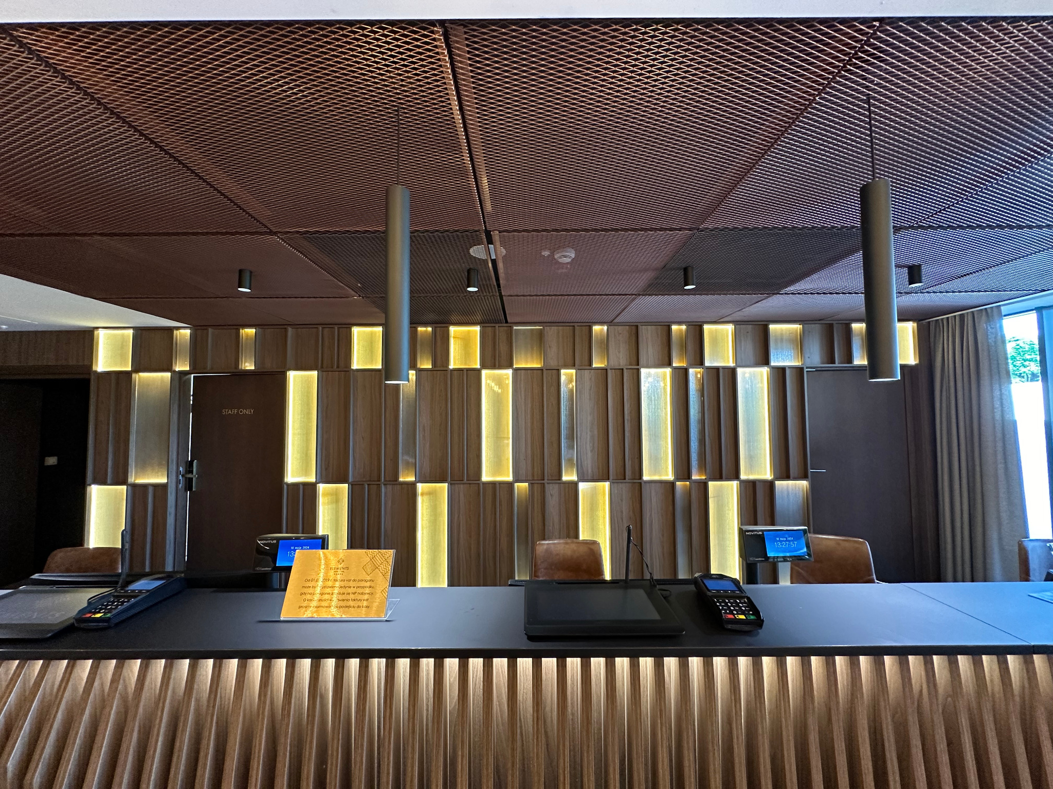 Recepcja hotelowa z drewnianą okładziną ścienną z podświetleniem, ciemnym blatem, kasami fiskalnymi i metalową kratką na suficie, oświetlona cylindrycznymi lampami wiszącymi.
