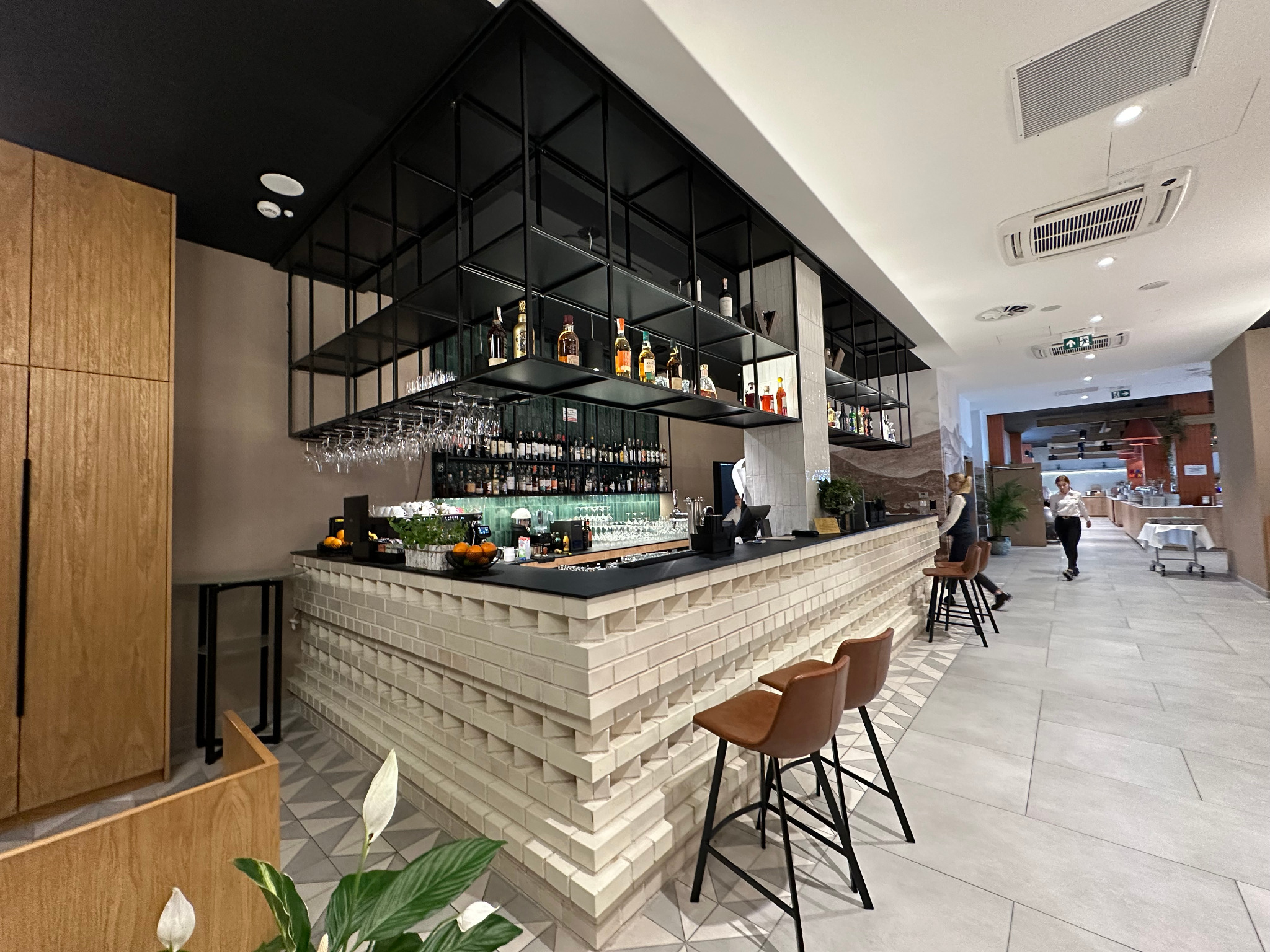 Nowoczesny bar z dekoracyjnym wykończeniem z cegieł, czarnym blatem i metalowymi półkami z alkoholem, widok na przestronne wnętrze restauracji z personelem.