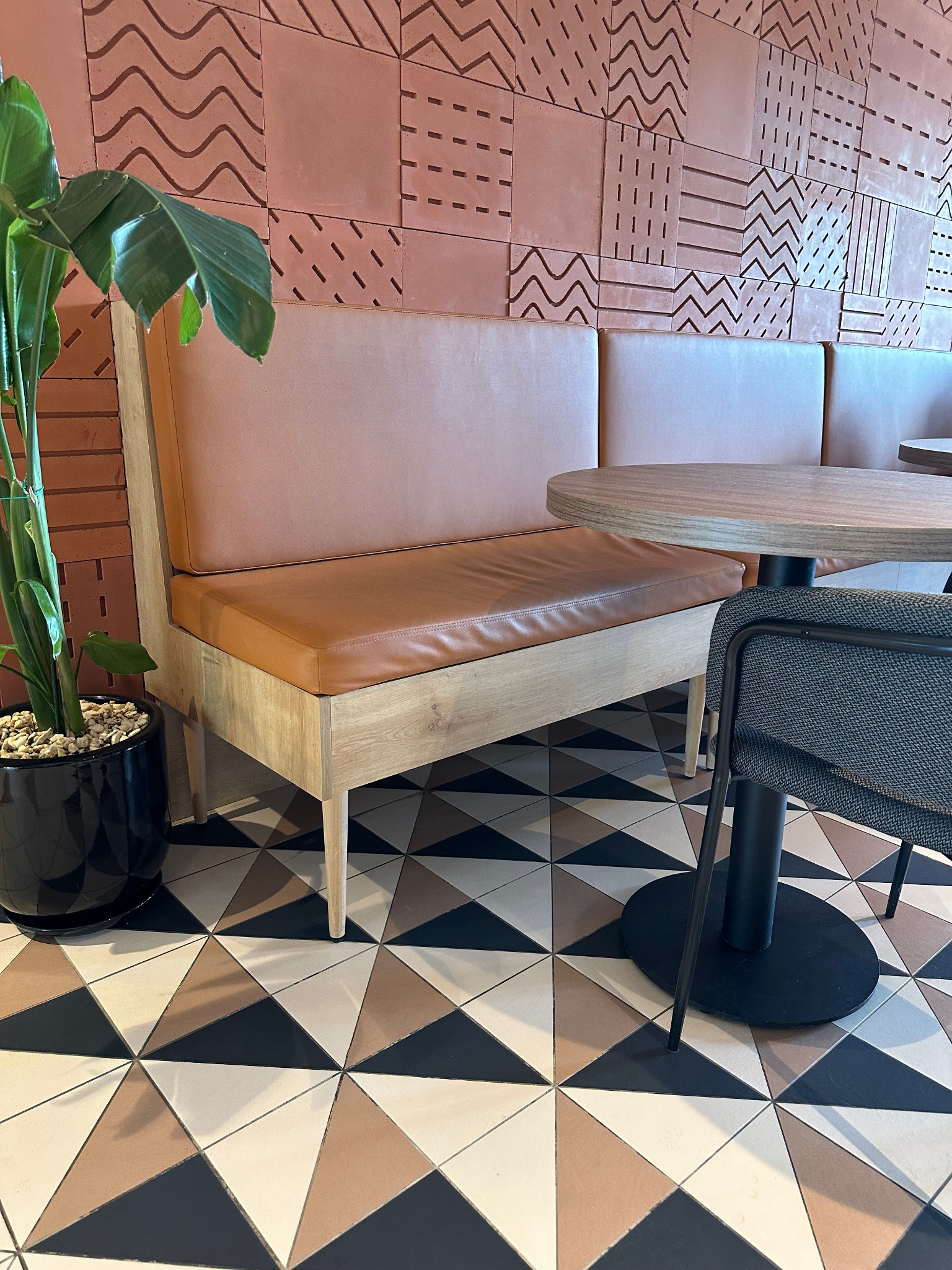 Wnętrze kawiarni z drewnianą ławką z brązowym siedziskiem, okrągłym stołem i krzesłem na tle ściany z dekoracyjnych płytek oraz wzorzystej podłogi z płytek.