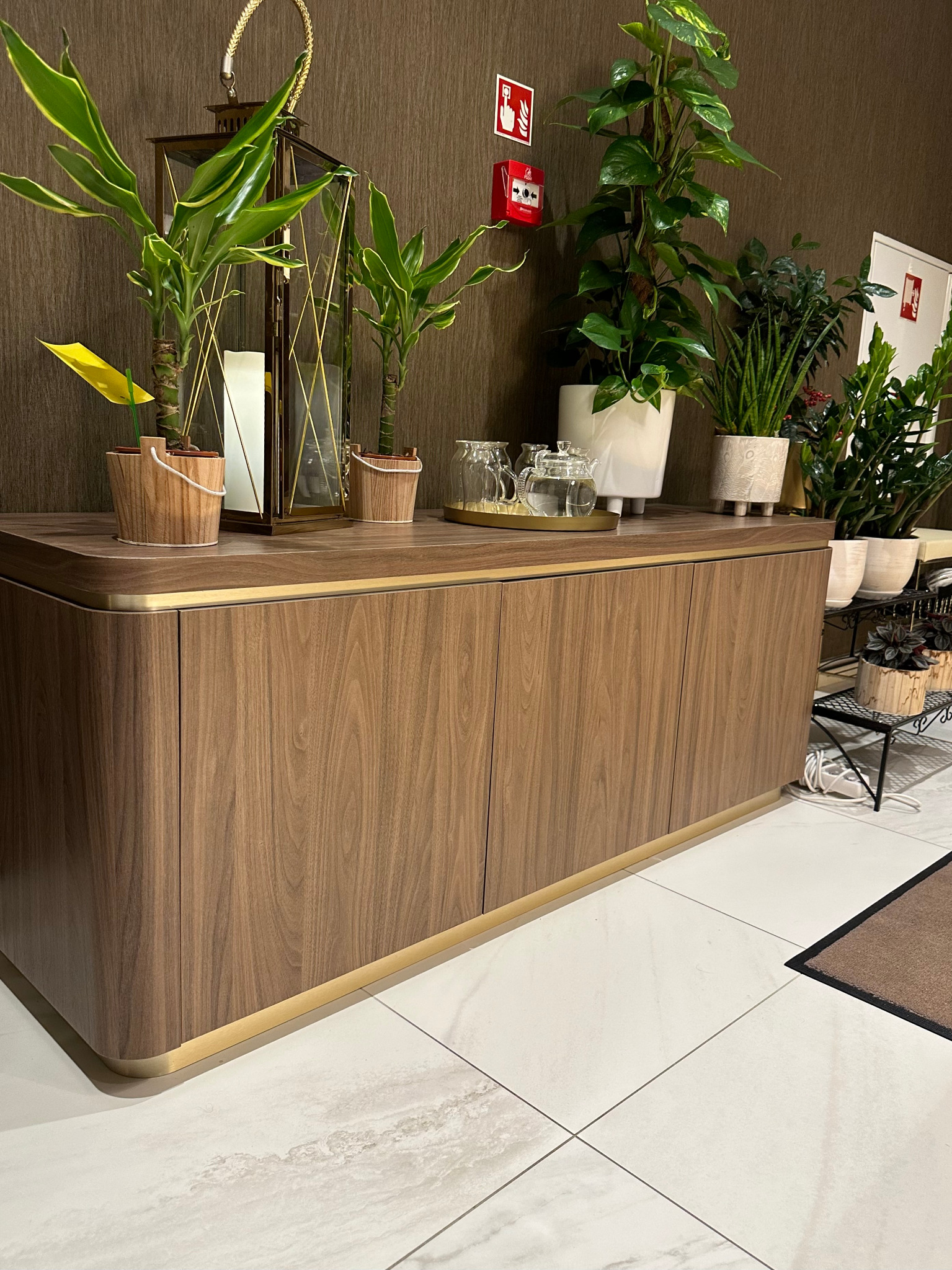 Drewniana komoda z zaokrąglonymi krawędziami i złotymi akcentami, udekorowana roślinami doniczkowymi, lampionem i szklanym serwisem do herbaty, ustawiona na białej marmurowej podłodze.