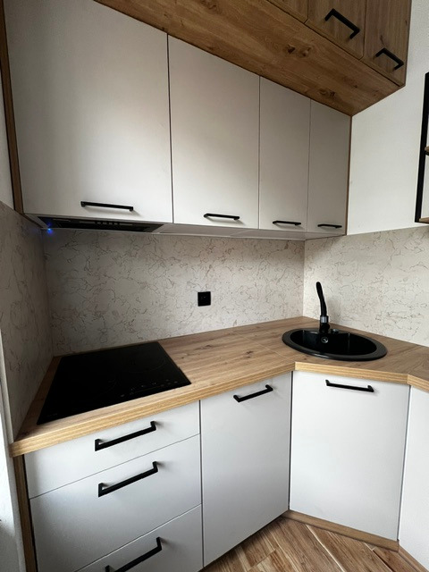 Nowoczesna kuchnia z białymi szafkami z czarnymi uchwytami, drewnianym blatem i czarnym zlewem owalnym, płyta indukcyjna w rogu blatu, widoczne szafki górne z drewnianym wykończeniem na górze.