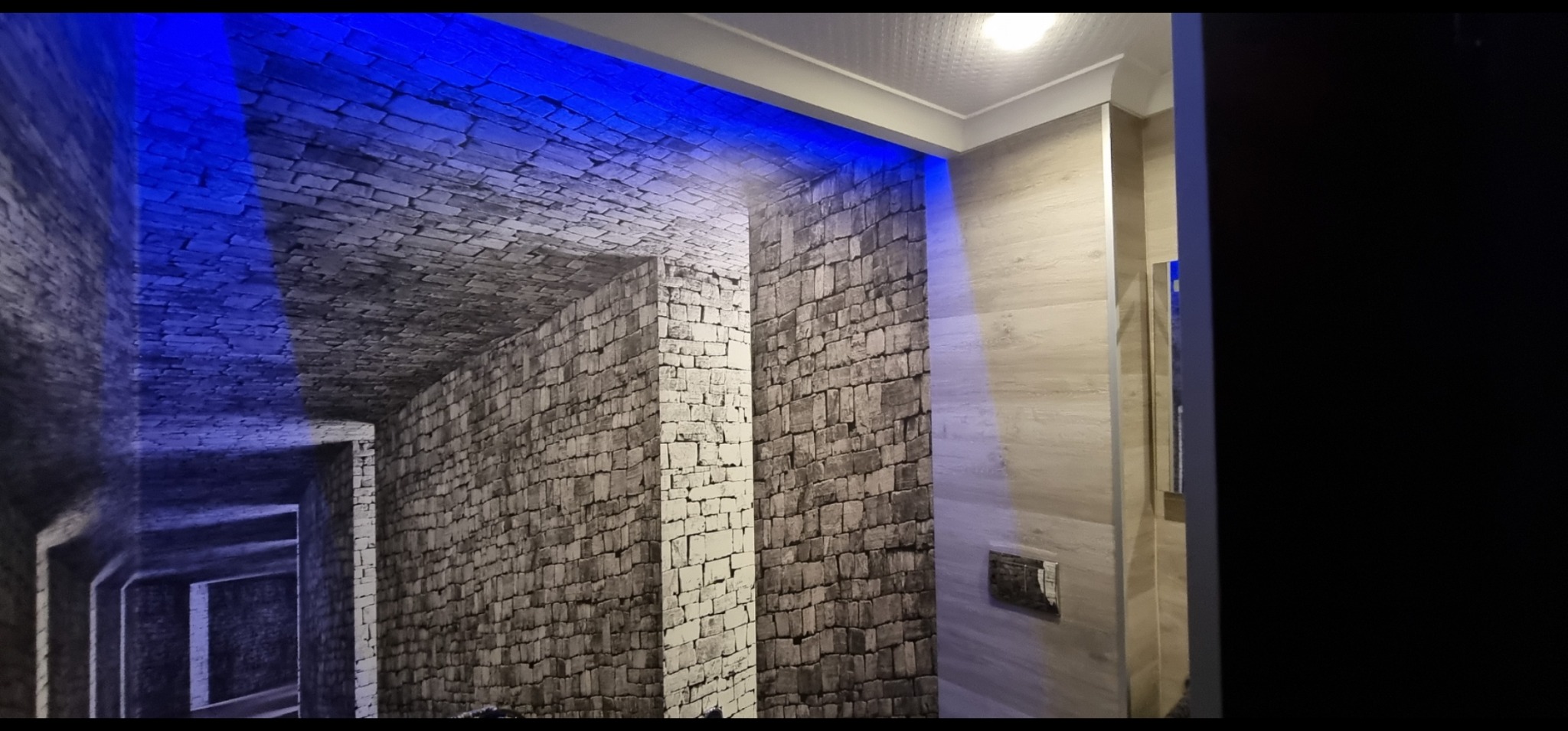 Ściana z fototapetą imitującą kamienny mur, podświetlona niebieskim światłem LED, sąsiadująca z jasną, wykończoną ścianą. Widoczny fragment łazienki.