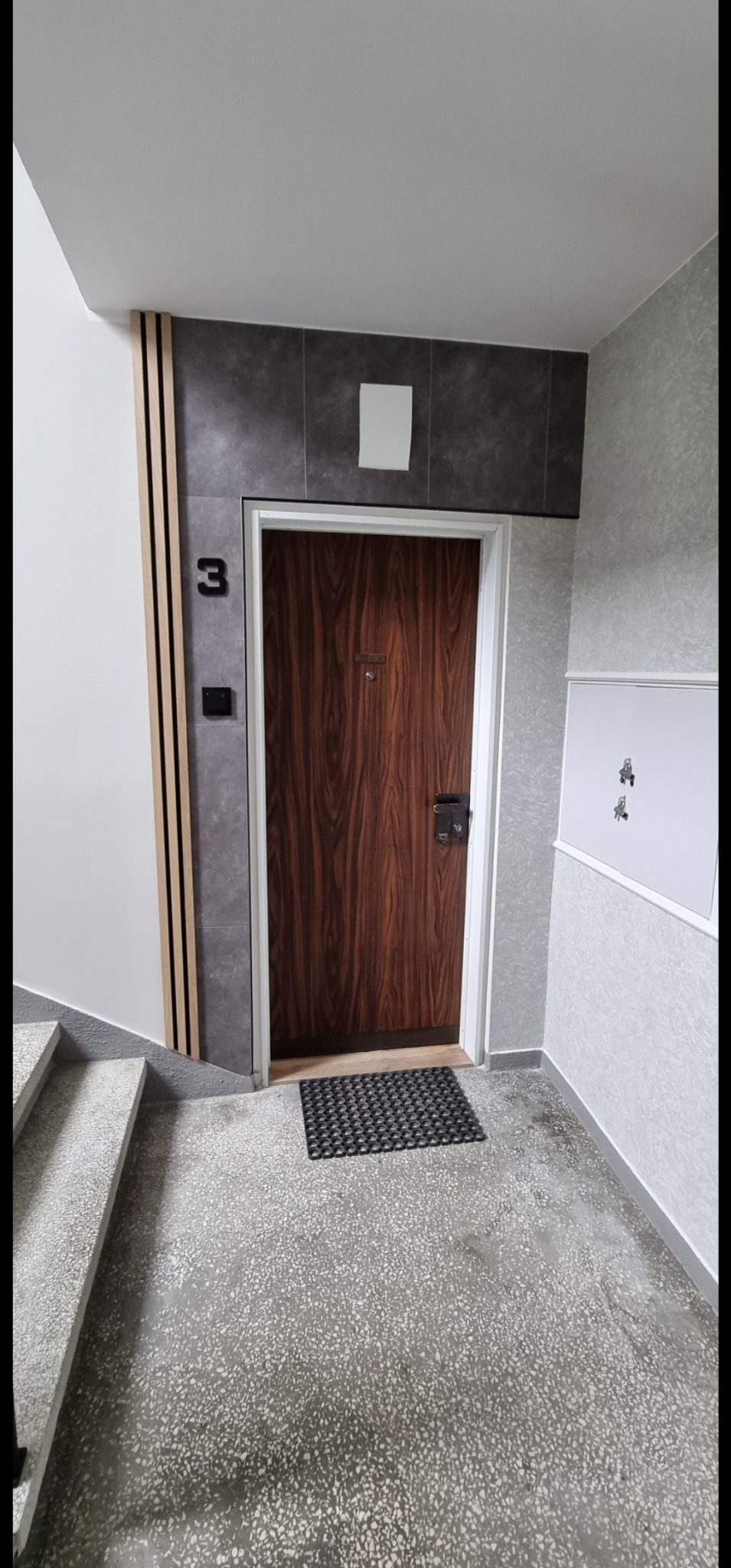 Wykończone wnętrze klatki schodowej: drewniane drzwi wejściowe nr 3, szara ściana z płytkami i drewnianymi panelami, oraz podłoga z lastryko. Nowoczesny design.