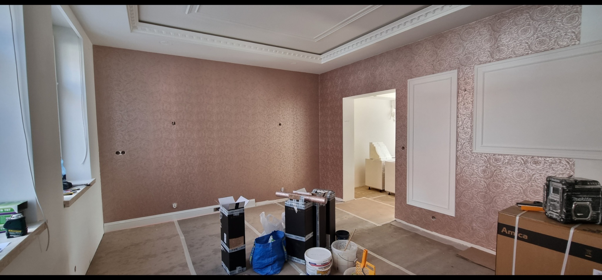 Pokój po tapetowaniu z dekoracyjną tapetą w odcieniach różu, widoczne narzędzia pracy na podłodze, listwy przysufitowe i ozdobne ramy na ścianach.