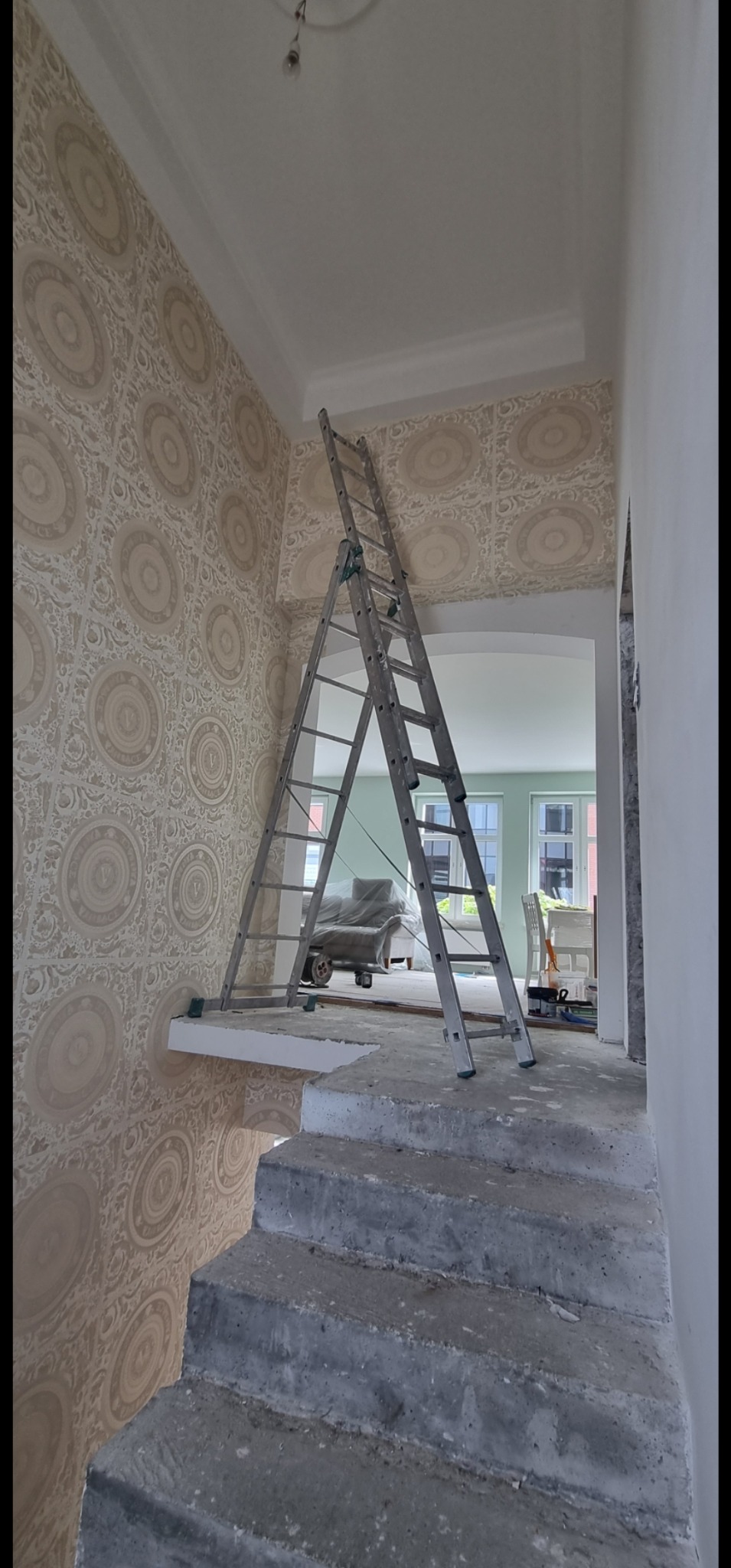 Schody z drabina na tle ściany pokrytej beżową tapetą w ornamenty. Widok na wnętrze z oknami. Prace wykończeniowe w trakcie, widoczne surowe schody.