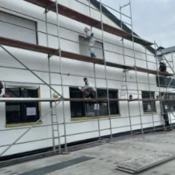 Trzech pracowników w kombinezonach malarskich na rusztowaniu maluje elewację budynku z zamkniętymi roletami okiennymi.