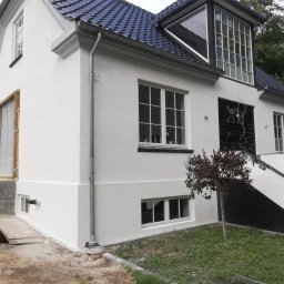 Remonty i ogrodnictwo - Biała fasada domu z ciemnym dachem i stolarką okienną, fragment okna zabezpieczony folią, widoczne ślady prac ziemnych wokół budynku.