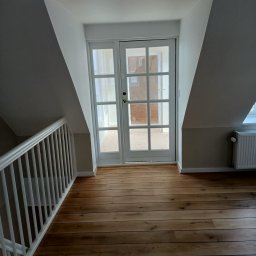 Remonty i ogrodnictwo - Poddasze po renowacji z drewnianą podłogą, białymi drzwiami balkonowymi i grzejnikiem. Widoczna biała balustrada schodów.