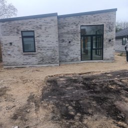 Remonty i ogrodnictwo - Nowoczesny dom z szarej cegły z czarnymi ramami okiennymi i drzwiowymi, teren wokół w trakcie prac ziemnych.