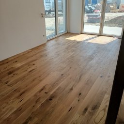 Remonty i ogrodnictwo - Nowo położona, jasna, drewniana podłoga z widocznymi sękami i naturalnym usłojeniem w pustym pomieszczeniu z dużymi oknami balkonowymi, przez które widać teren budowy.