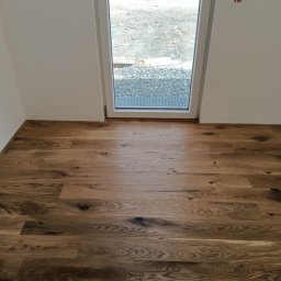 Remonty i ogrodnictwo - Ułożona drewniana podłoga z widocznymi słojami i sękami, z fragmentem okna wychodzącego na plac budowy z paletami cegieł.