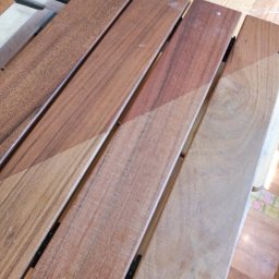 Remonty i ogrodnictwo - Cztery drewniane deski połączone czarnymi metalowymi elementami, z widoczną różnicą w odcieniach brązu po malowaniu lub lakierowaniu, ułożone na drewnianym stole, z butelką w tle.