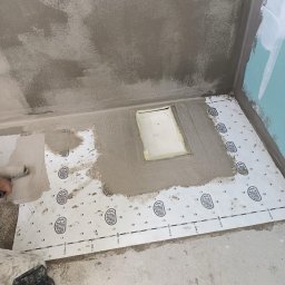 Remonty i ogrodnictwo - Przygotowanie do hydroizolacji podłogi w łazience w Gorzowie Wielkopolskim: aplikacja masy uszczelniającej wałkiem na folii w płynie marki Ardex, widoczne surowe ściany i wnęka odpływu liniowego.