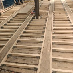 Remonty i ogrodnictwo - Konstrukcja drewniana podłogi z widocznymi legarami i wspornikami kątowymi, przygotowywana do dalszych prac wykończeniowych, z pracownikami w tle.