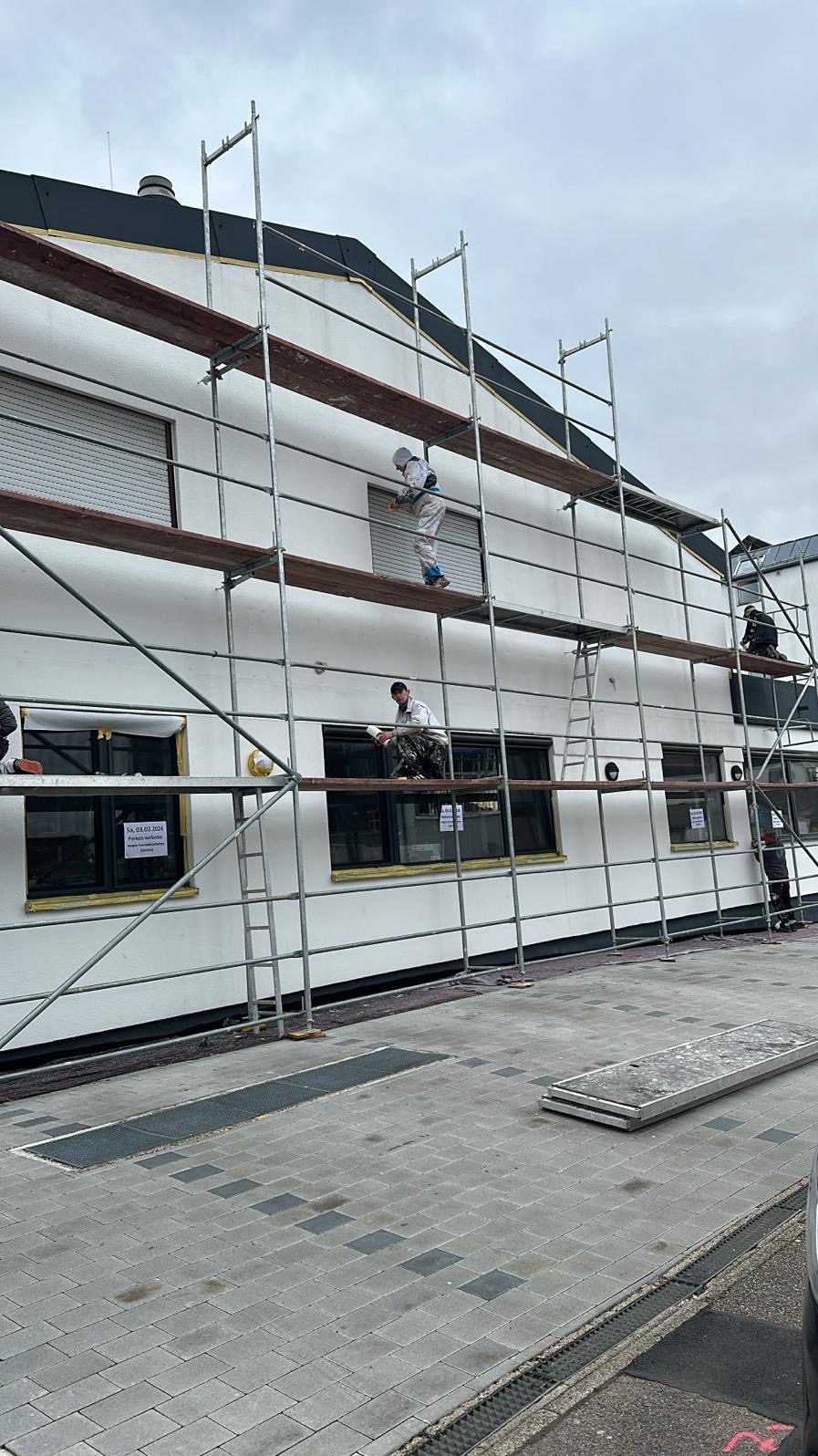 Trzech pracowników w kombinezonach malarskich na rusztowaniu maluje elewację budynku z zamkniętymi roletami okiennymi.