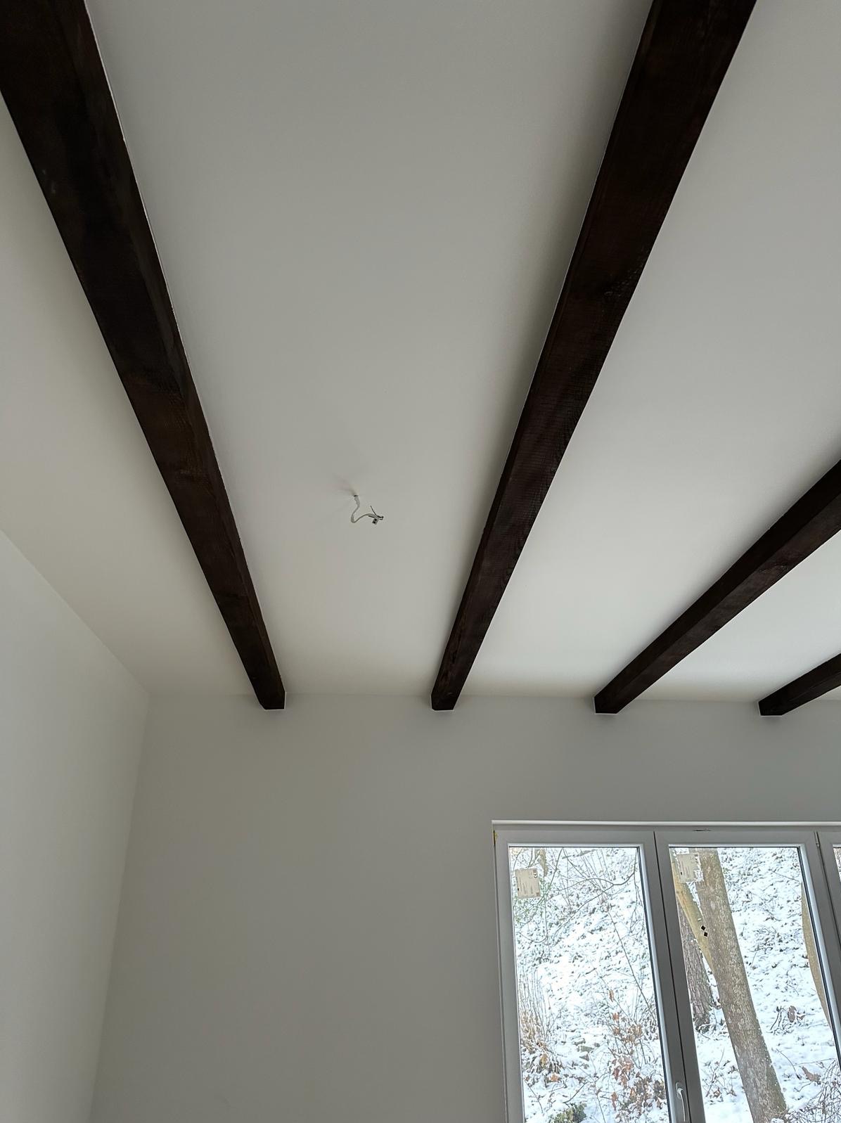 Biały sufit z drewnianymi belkami stropowymi w nowo wykończonym pokoju, widoczne okno z zimowym krajobrazem.