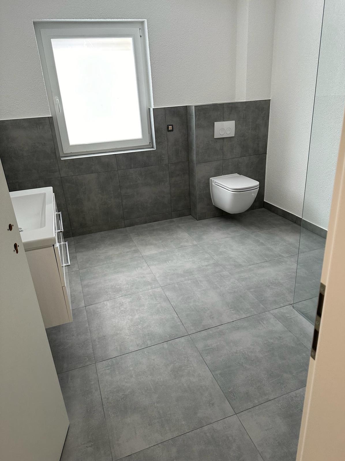 Nowoczesna łazienka z szarymi płytkami, podwieszaną toaletą i umywalką z szafką, oświetlona naturalnym światłem z okna.