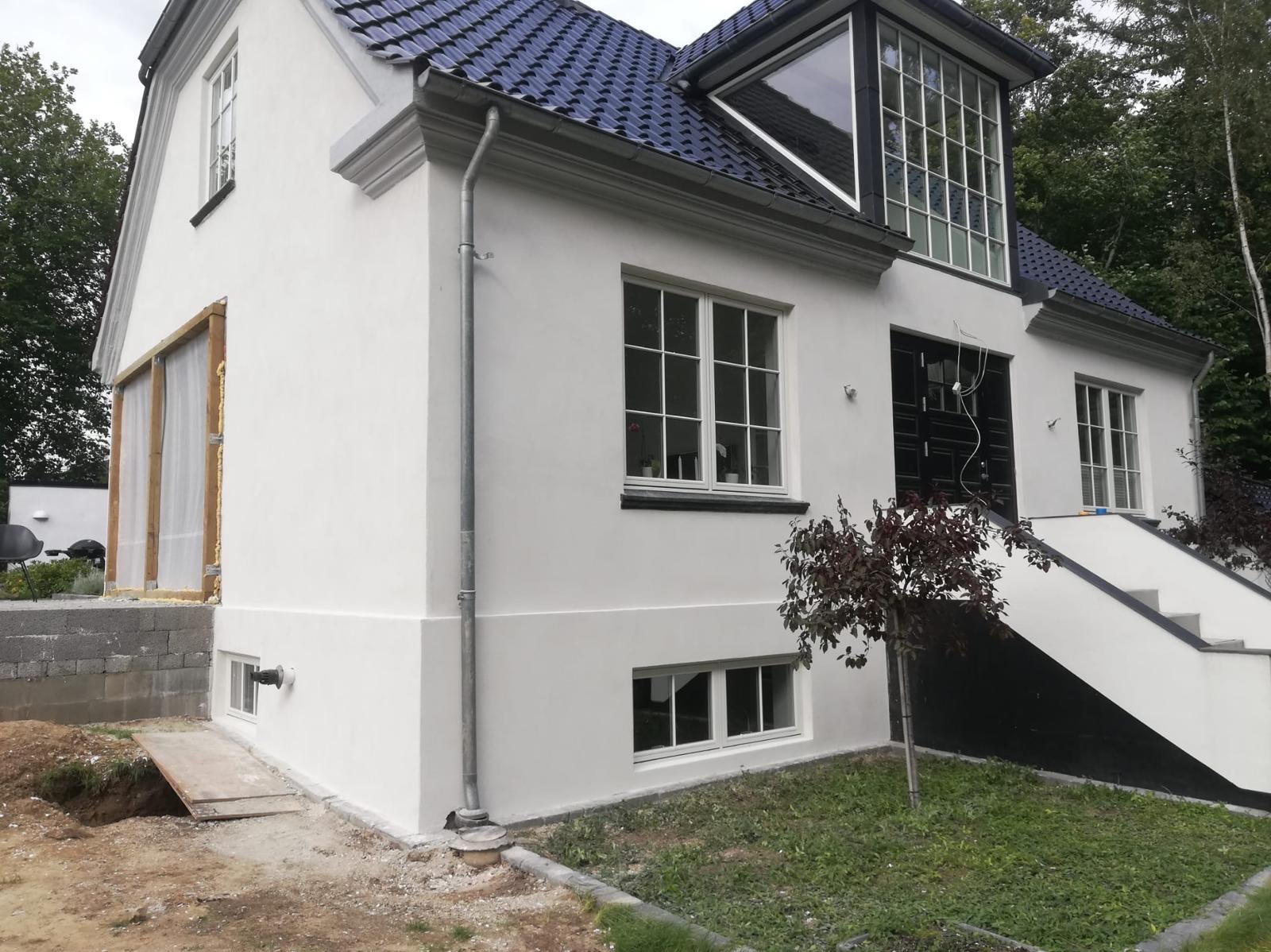Biała fasada domu z ciemnym dachem i stolarką okienną, fragment okna zabezpieczony folią, widoczne ślady prac ziemnych wokół budynku.