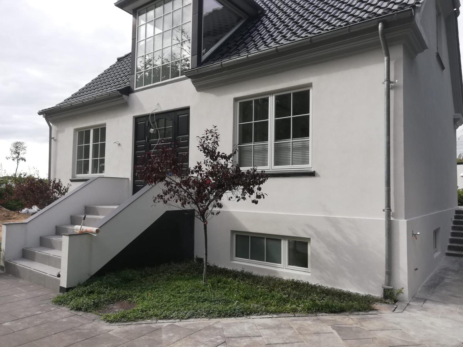 Elewacja domu jednorodzinnego po renowacji, świeżo pomalowane ściany na biało, widoczne nowe schody wejściowe i odnowiona stolarka okienna.