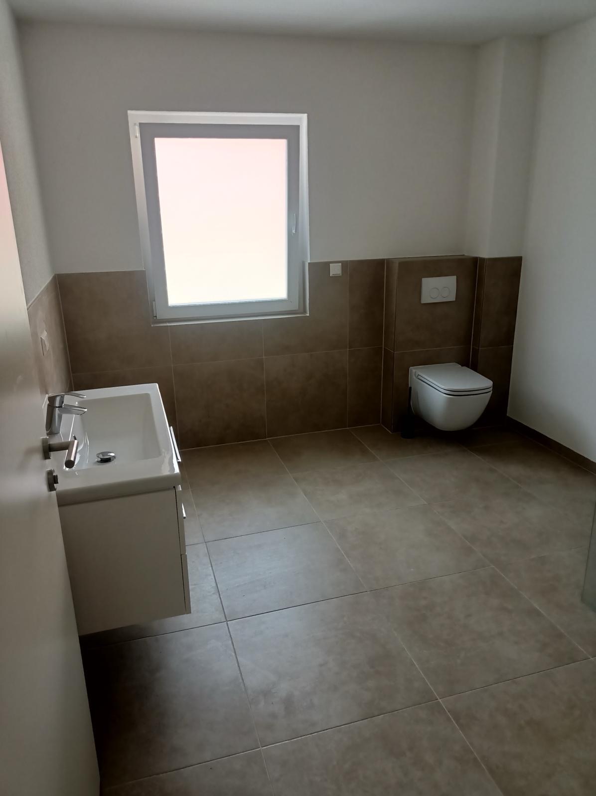 Nowoczesna łazienka z brązowymi płytkami, białą umywalką z szafką i wiszącą toaletą, oświetlona przez okno z białymi ramami.