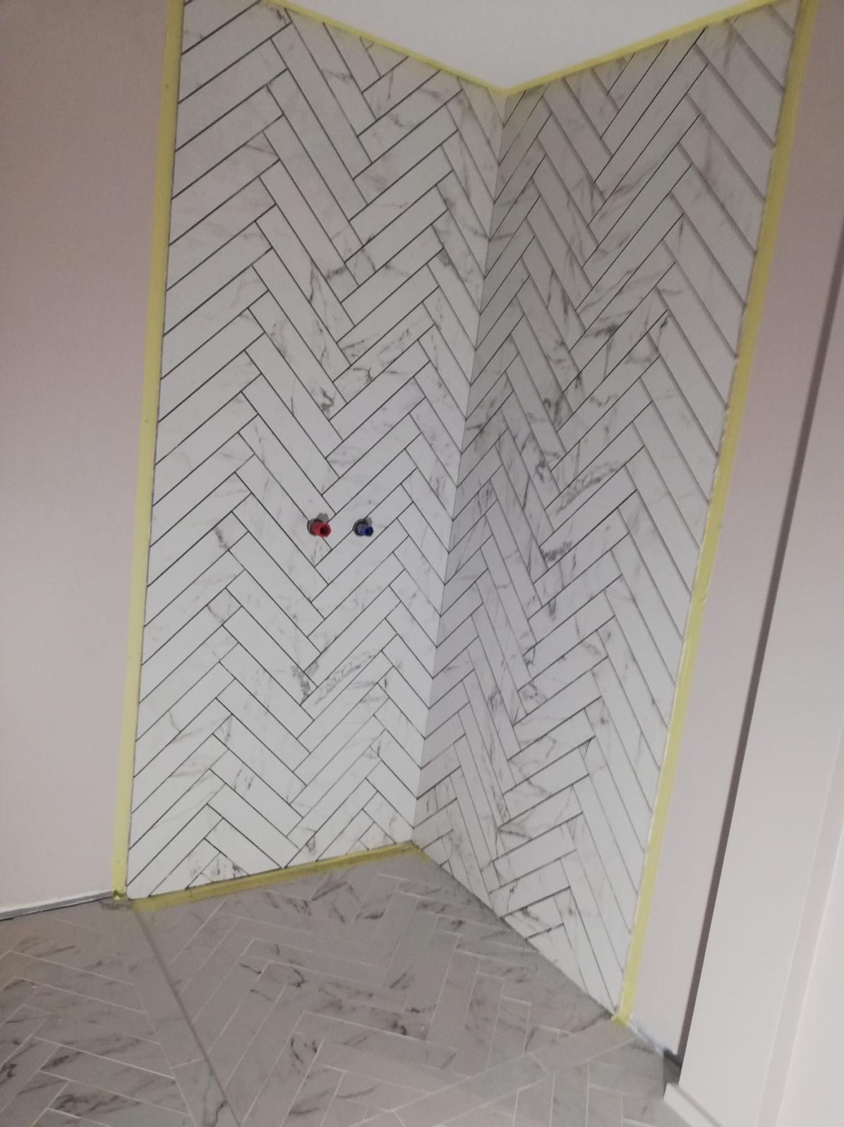 Ściana i podłoga wyłożone białymi płytkami imitującymi marmur w jodełkę, zabezpieczone żółtą taśmą malarską, widoczne przyłącza wodne.