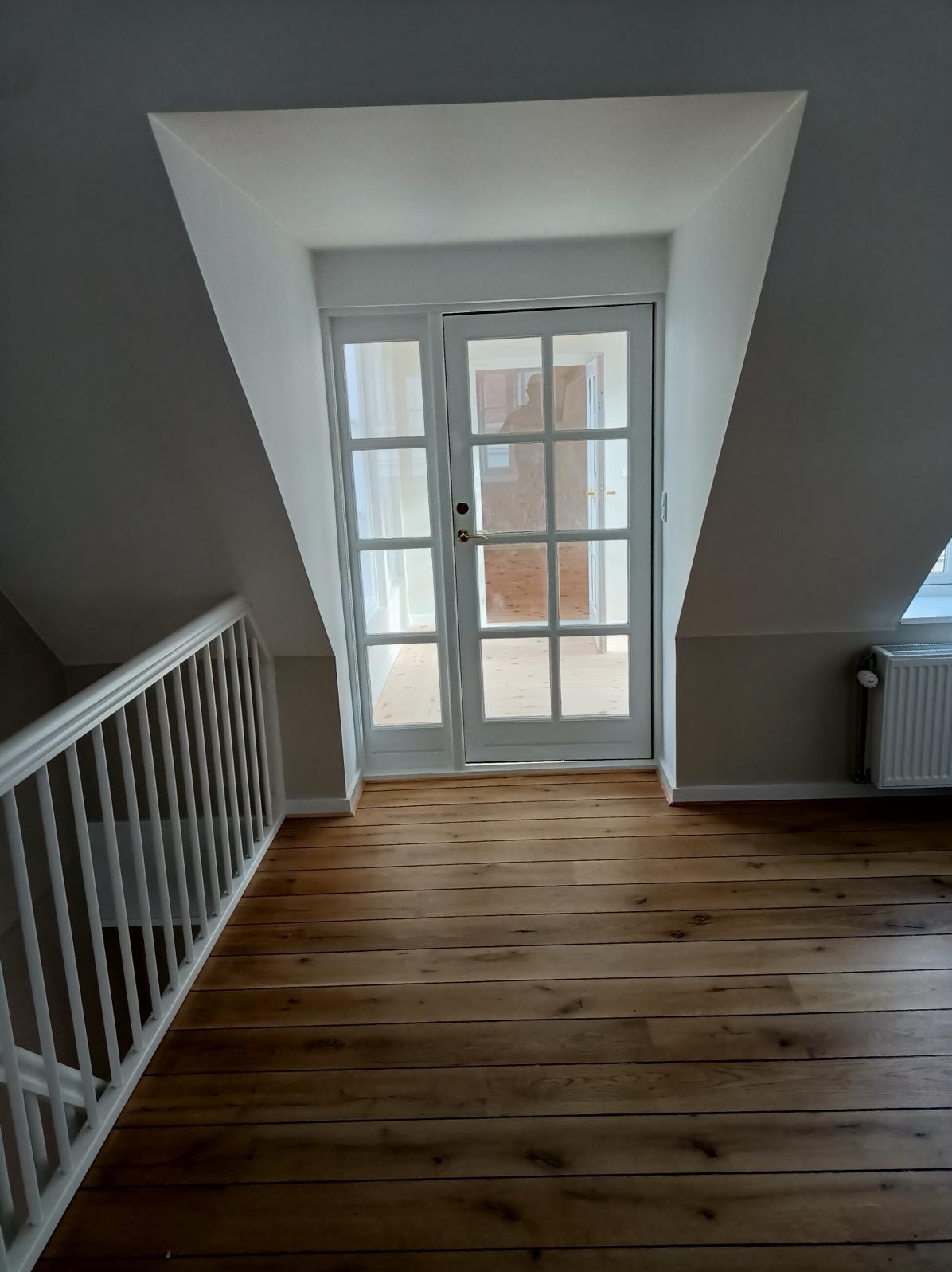 Poddasze po renowacji z drewnianą podłogą, białymi drzwiami balkonowymi i grzejnikiem. Widoczna biała balustrada schodów.